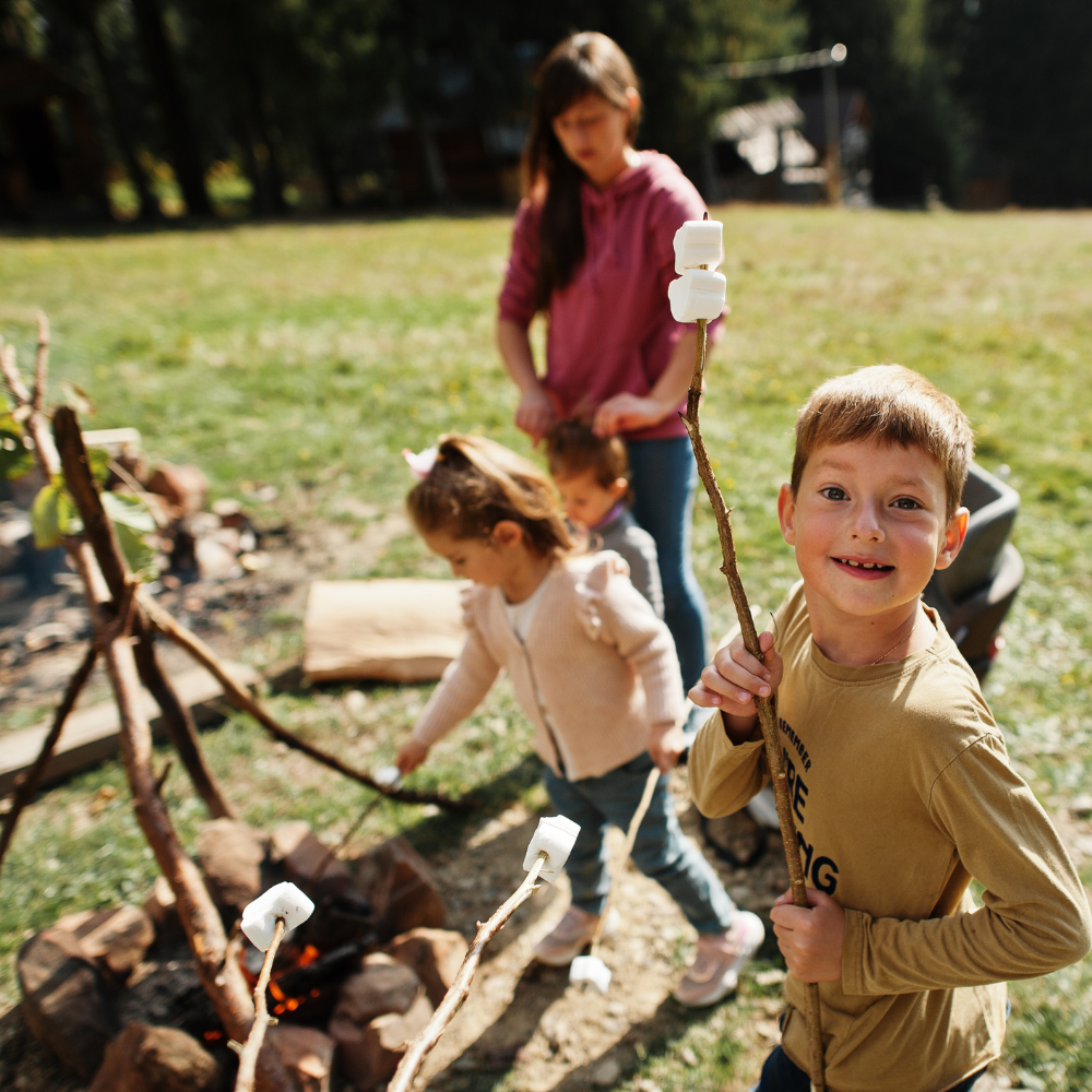 Young boy holding a stick with marshmallows. Behind him is a makeshift fire with a young girl and adult roasting marshmallows.