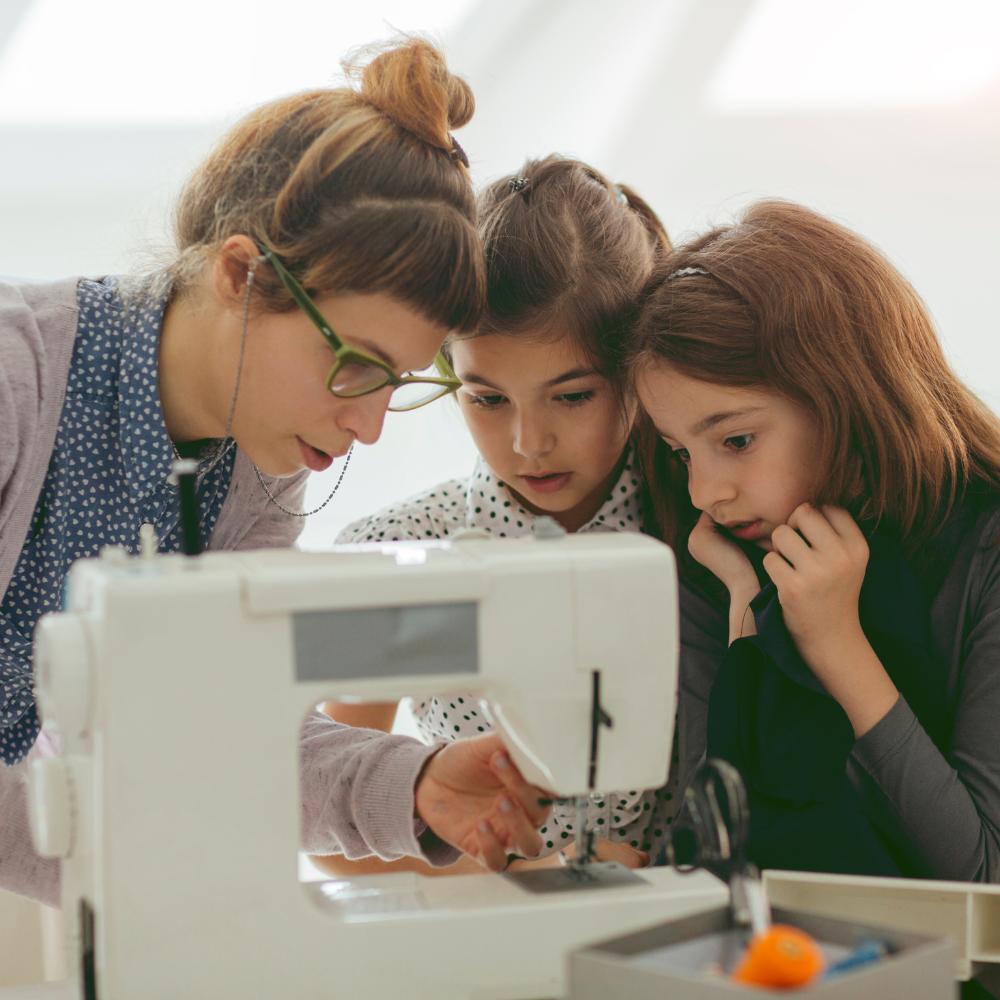 Two girls and a woman work together on a garment using a sewing machine